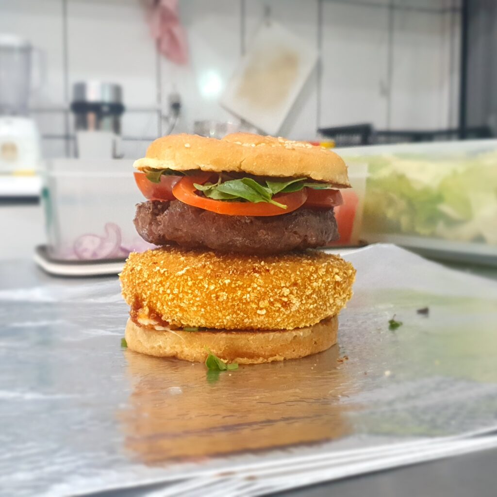 imagem do bog 4 queijos que possui um hamburguer de carne, pão sem glúten e sem lacteos, hamburguer de 4 queijos sem glúten, tomate orégano e manjericão. Está em uma mesa com a cozinha de fundo.