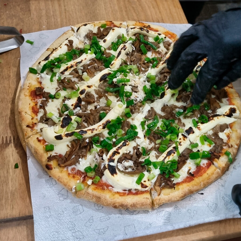 Pizza sem glúten, recheio de mussarela, shimeji, cream cheese e cebolinha e a mão do operador.